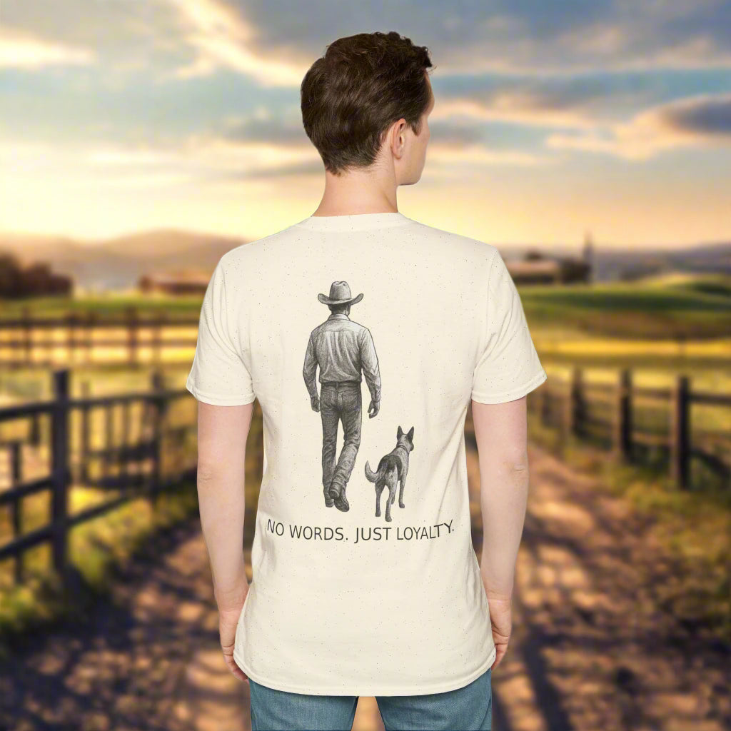 Cowboy wearing a light-colored long-sleeve shirt and jeans, walking side by side with a dog. The back of the t-shirt features detailed artwork of the cowboy and dog with the slogan "NO WORDS. JUST LOYALTY." printed below. Front chest logo shows "Adopted & Adored Apparel."