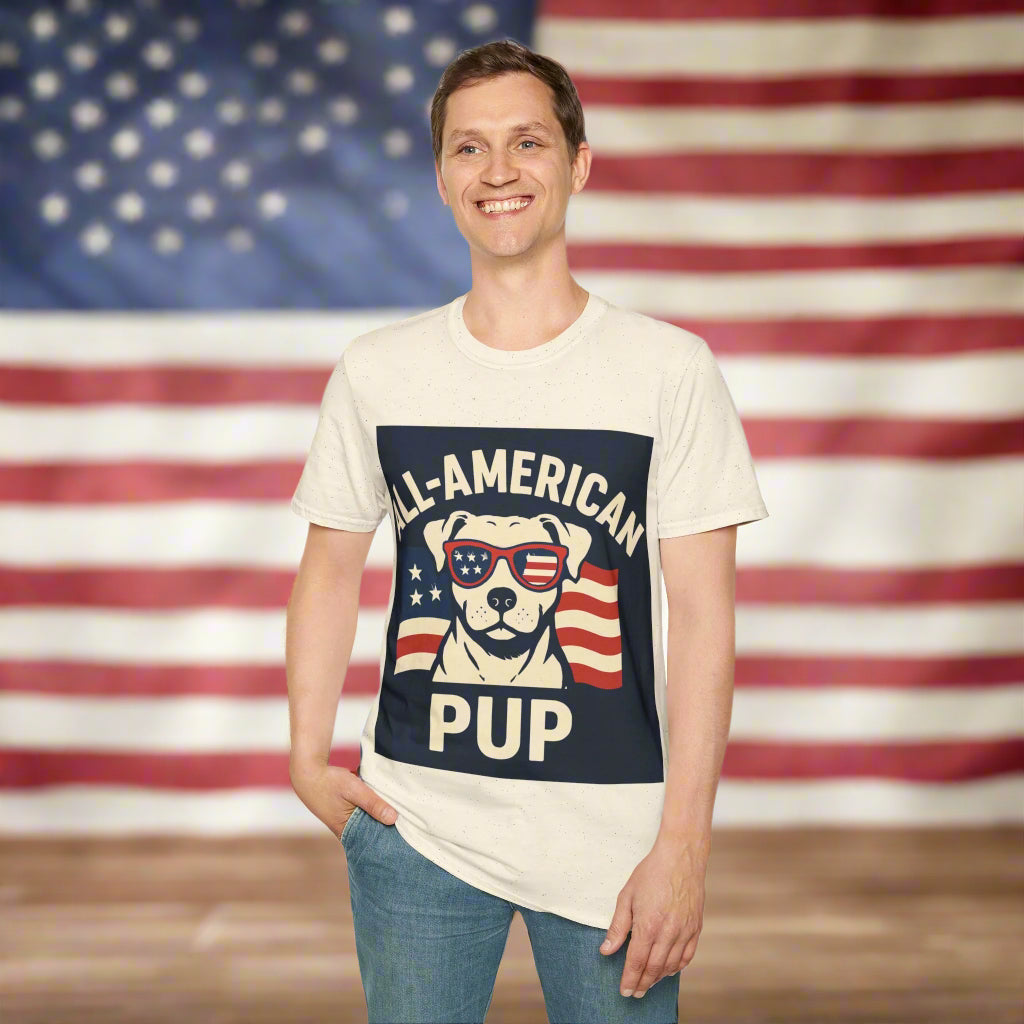 Person wearing a tan t-shirt that says “All-American Pup,” standing in front of a faded American flag background. The shirt design is bold and clearly visible.