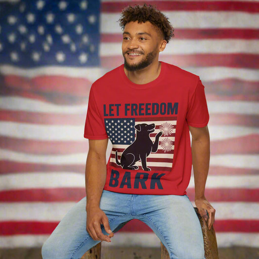 Man wearing a red t-shirt that says “Let Freedom Bark” with blue lettering, featuring a black dog and flag-themed graphic. He is sitting on a pedestal in an urban city background, facing forward.

