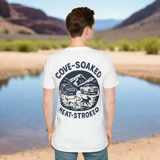 Man wearing white t-shirt with navy blue back print standing by a desert lake. Shirt features dogs running in water, 4x4 trucks, mountains, and “Cove-Soaked Heat-Stroked” text.
