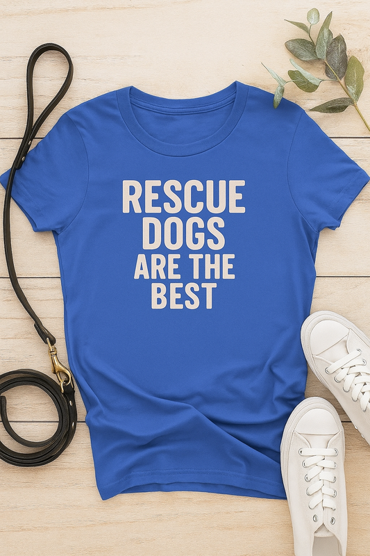 Royal blue women's t-shirt laid flat on a light wood background, featuring the phrase "Rescue Dogs Are the Best" in distressed cream text. Styled with a black leather dog leash, white canvas sneakers, and eucalyptus leaves for a soft, dog-lover aesthetic.