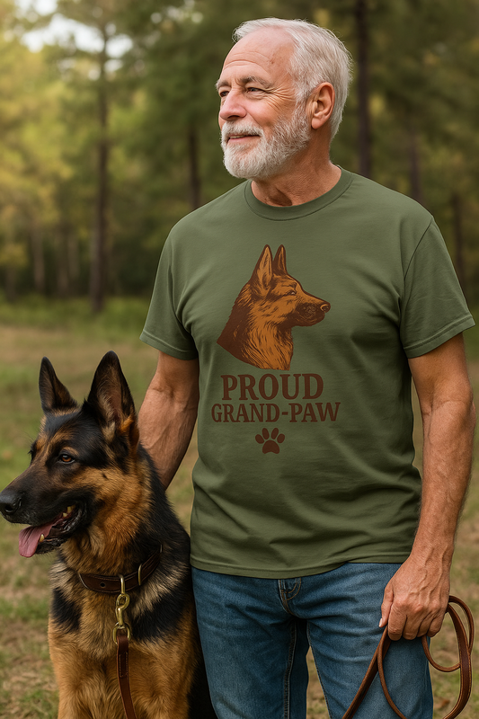 Senior man holding a leash beside a German Shepherd in a wooded area, wearing an olive green "Proud Grand-Paw" t-shirt with a rustic German Shepherd graphic and distressed paw print.