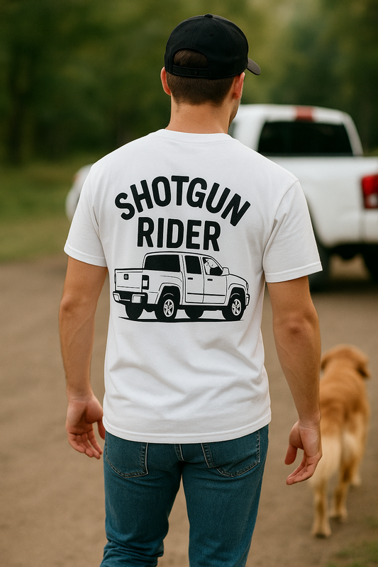 Young man in his 20s wearing a white "Shotgun Rider" t-shirt with a bold pickup truck graphic on the back. He walks on a dirt path with a golden retriever beside him, approaching a white truck in the blurred background. The scene evokes a laid-back, outdoorsy lifestyle with a strong dog-and-truck bond.