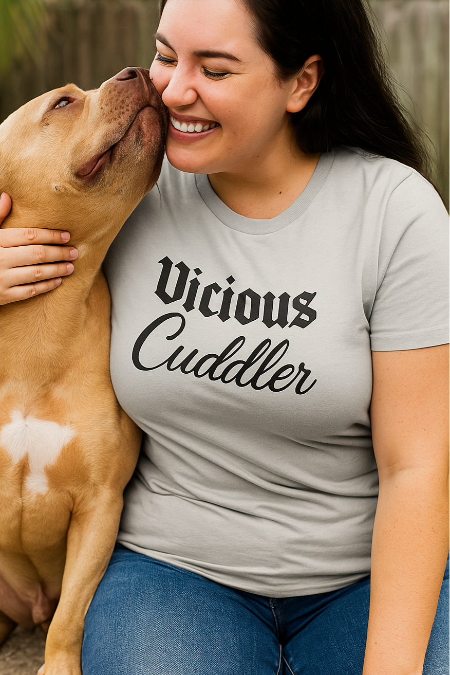 Smiling woman wearing a “Vicious Cuddler” t-shirt sits outdoors with her muscular tan pit bull, who affectionately leans in to nuzzle her face. The scene highlights their loving bond and the contrast between the bold shirt message and the dog’s gentle behavior.