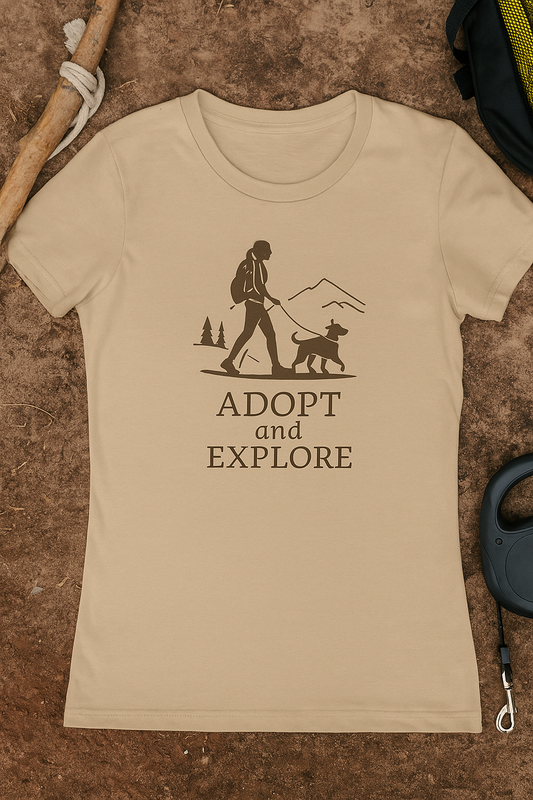 Beige women's t-shirt laid flat on rugged ground, featuring a brown “Adopt and Explore” graphic of a female hiker walking a dog with mountain and tree silhouettes in the background. Surrounded by a wooden hiking stick, coiled dog leash, and backpack gear.